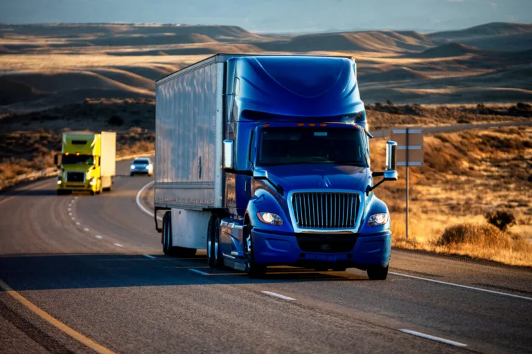A semi-truck with a blue cab and a white trailer drives on a highway at sunset. Another truck is visible behind it.