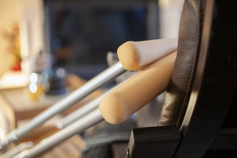 A pair of crutches resting against the arm of a brown leather chair, with a blurred background.