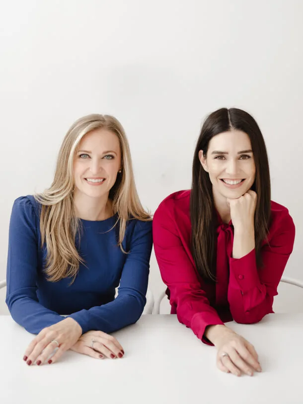 A photo of two smiling women, one in a blue dress and the other in a red blouse, seated at a white table.
